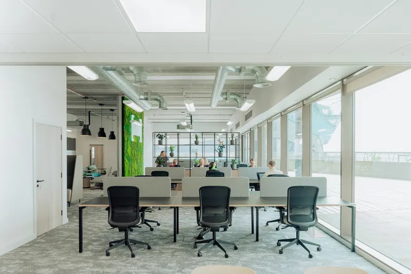Bright open-plan workplace with desks, glass meeting pods and a planted divider, people working at laptops